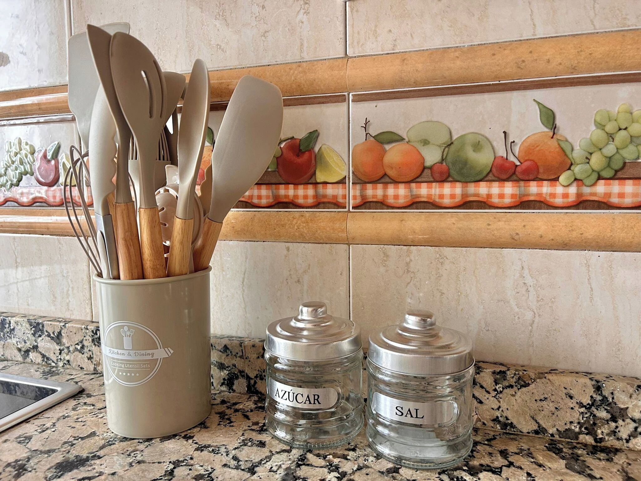 A kitchen counter with a utensil holder, two glass jars labeled 'AZÚCAR' and 'SAL', and a decorative backsplash with fruit designs.