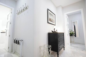 Un couloir aux murs blancs, une commode noire, un tableau encadré et un porte-manteau avec des manteaux.