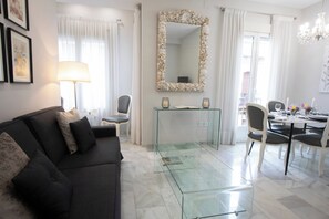 Un salon moderne avec une table basse en verre, un canapé foncé, un coin repas avec une table et des chaises blanches, et un grand miroir avec un cadre en forme de coquillage.