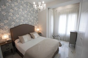 A bedroom with a bed, nightstands, a lamp, a chandelier, a window with curtains, and a radiator. - La terraza de Alfonso XIII (Cordoba)