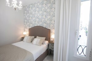 Ein Schlafzimmer mit einem Bett, zwei Nachttischen mit Lampen, einem blumigen Wandpaneel als Kopfteil und Blick auf die Straße durch ein Fenster.