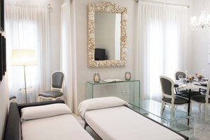 A hotel room with two beds, a glass table, a chair, a desk, and a mirror with a shell frame. - La terraza de Alfonso XIII (Cordoba)