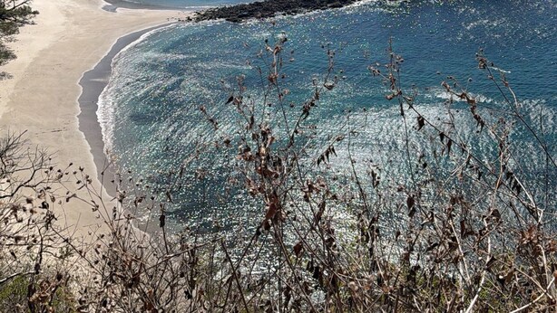 Eine Küstenlandschaft mit klarem Wasser, einem Sandstrand, Felsformationen und einem bergigen Hintergrund.
