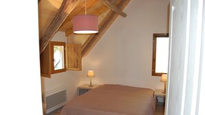 Un dormitorio con techo inclinado, una cama con colcha rosa, dos mesitas de noche con lámparas y un ventanal.