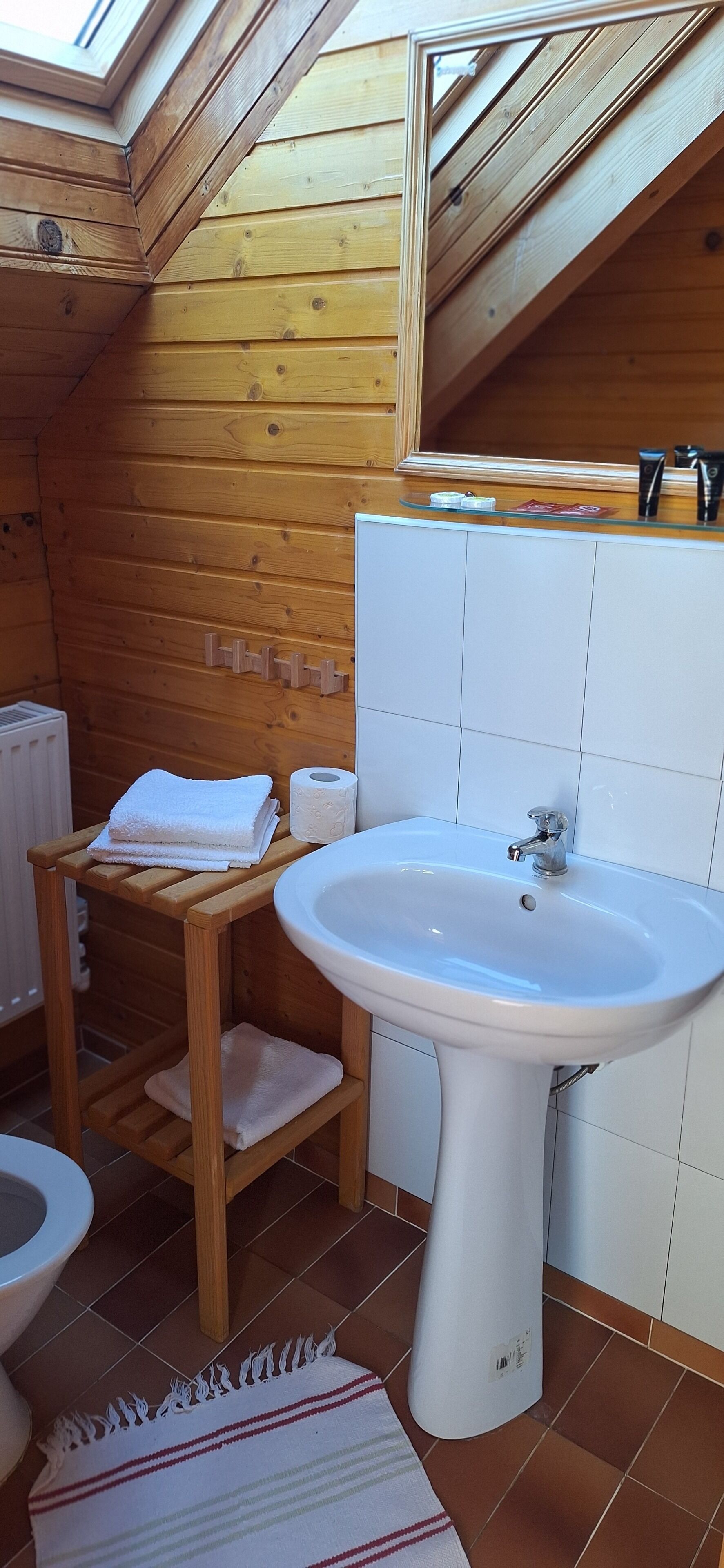 A wooden cabin bathroom with a skylight, a white sink, a wooden shelf with towels, and a tiled floor.