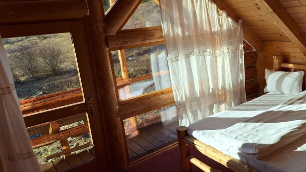 A wooden cabin room with a bed, a window with curtains, and a view of a forested landscape. - Chalet Rosehip Flowers 4-6 Traveling (Rasnov)