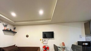 A living room with a brown sofa, a glass table, a red fan, a flat-screen TV, and a small shelf with decorative items.