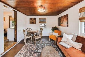 A cozy living area with a wooden ceiling, a round dining table with chairs, a sofa with cushions, and a large window with blinds. - The Mystic Inn | Wine and Cheese Shop Downstairs | Downtown LG (Lake Geneva)