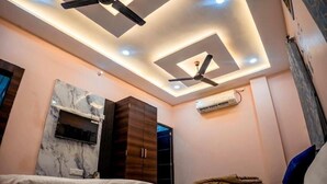 A bedroom with a bed, a wardrobe, a ceiling fan, recessed lighting, and a TV mounted on the wall. - Shiv Darshan P Guest House (Varanasi)