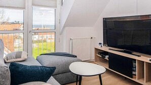 A modern living room with a large TV, a sofa, a small table, and a view through large windows. - 4 Star Holiday Home in Hejls (Hejls)