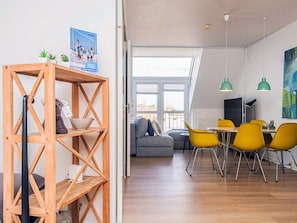 Un intérieur moderne avec une étagère en bois, un coin repas avec des chaises jaunes, un canapé gris et une grande fenêtre.