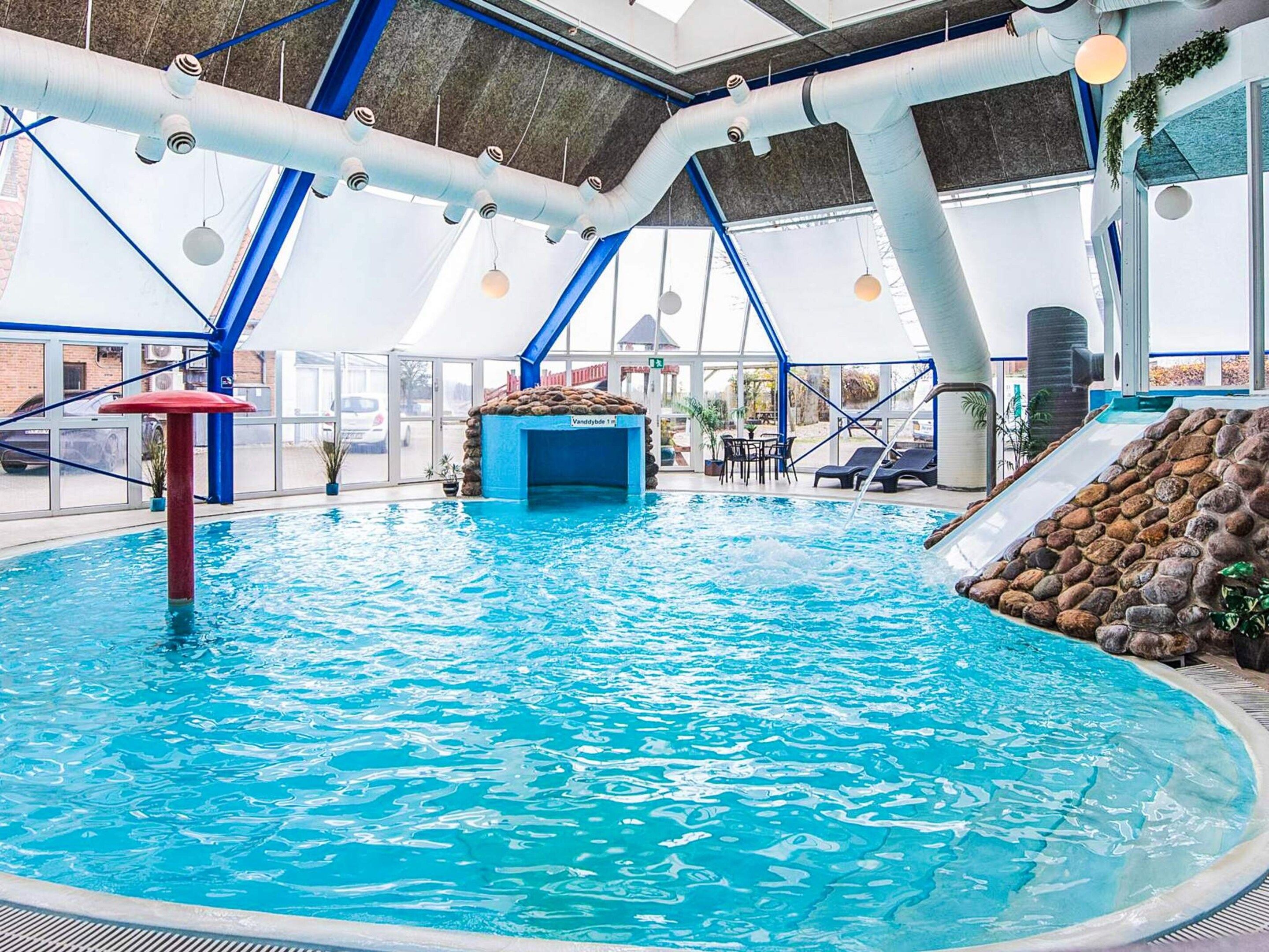 An indoor swimming pool with a clear view of the ceiling structure, a water slide, and a seating area with tables and chairs.