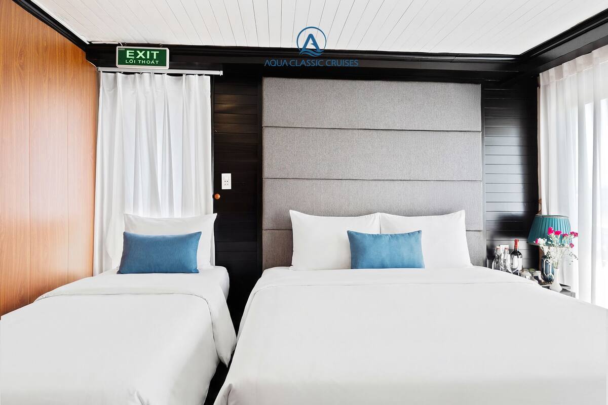 Hotellihuone, jossa on kaksi sänkyä, opastekyltti, joka osoittaa uloskäynnin, ja Aqua Classic Cruises -yhtiön logo.
