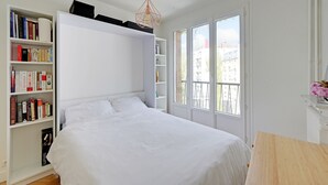 Ein Schlafzimmer mit einem Bett, weißen Wänden, einem Bücherregal, einem Fenster und einer Pendelleuchte.