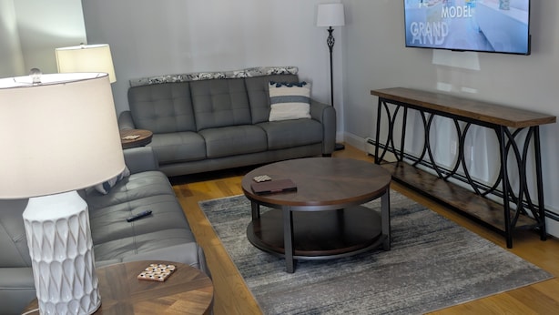Una sala de estar moderna con un sofá gris, una mesa redonda de centro, una mesita auxiliar de madera, una lámpara de pie y un televisor de pantalla plana montado en la pared.