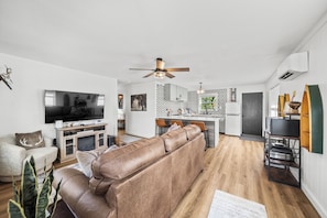 A modern living room with a large flat-screen TV, a leather sofa, a dining area, and a kitchen with a patterned backsplash.