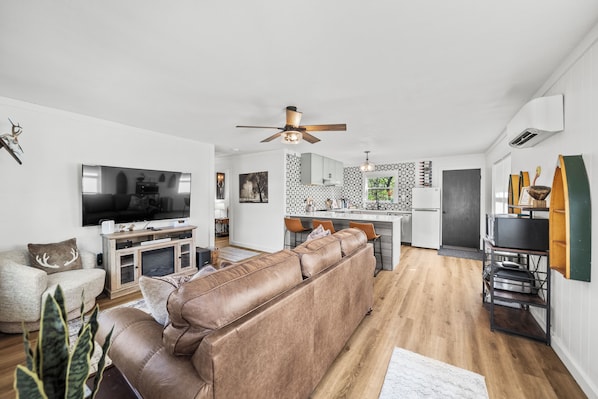 A modern living room with a large flat-screen TV, a leather sofa, a dining area, and a kitchen with a patterned backsplash.