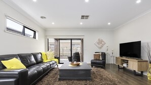 A modern living room with a black leather sofa, a dark coffee table, a TV stand, and a large window. - Comfort and Space in Lara's Family Friendly Home (Geelong)