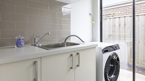 A kitchen with a built-in washing machine, a sink, and a window with a view of a wooden fence. - Comfort and Space in Lara's Family Friendly Home (Geelong)