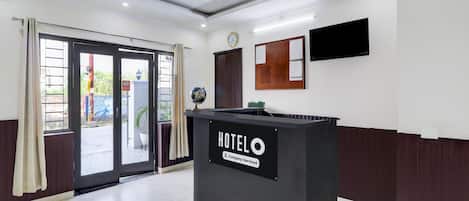 Zona de recepción de un hotel con un mostrador de recepción identificado como 'HOTELO Company-Serviced', un reloj, un televisor montado y un amplio ventanal con vistas al exterior.