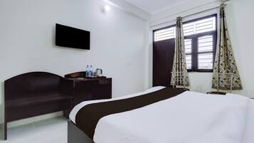 A hotel room with a bed, a desk with a flat-screen TV, a window with curtains, and a wall-mounted air conditioning unit.