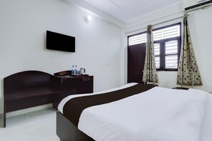 A hotel room with a bed, a desk with a flat-screen TV, a window with curtains, and a wall-mounted air conditioning unit. - Deluxe Benison Guest House (Dehradun)