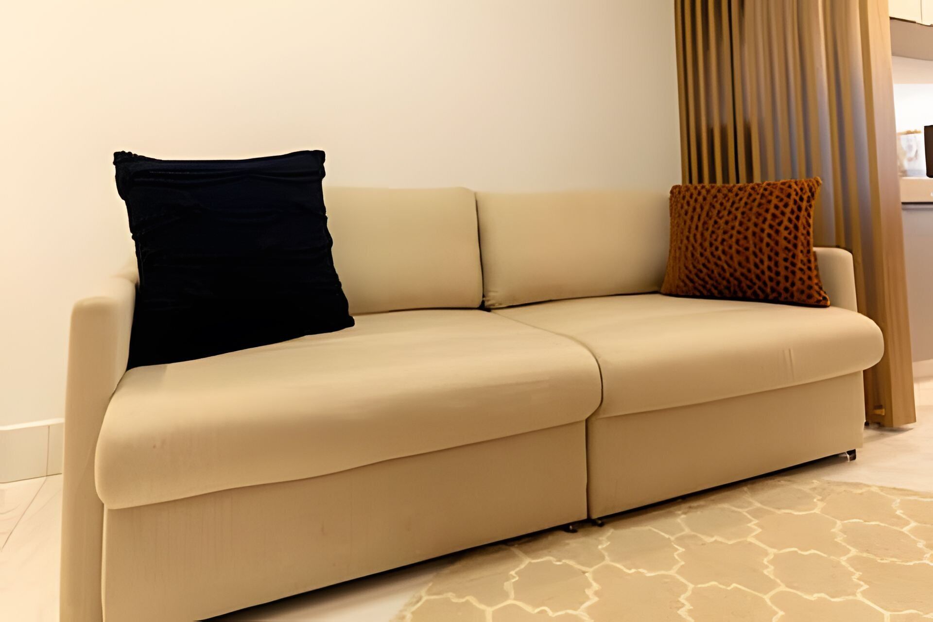 Un sofá color beige con dos almohadas, un tapiz estampado y una ventana con cortinas.