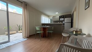 Un espacio habitable compacto con kitchenette, una zona de comedor con dos sillas, un sofá y un ventanal corredero que da a un área exterior.
