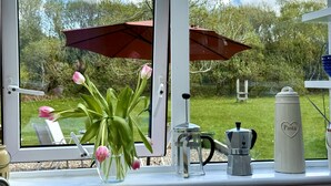 Una finestra con vista su un giardino, un tavolo con una macchina da caffè e un vaso con fiori.