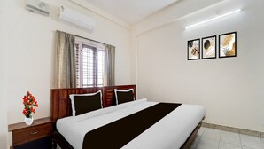 A hotel room with a bed, bedside table, wall-mounted air conditioner, window with curtains, and framed artwork.
