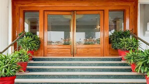 A wooden double door with glass panels, flanked by red potted plants, leading to a staircase. - Deluxe Room Subhash Nagar Royal Palazzo (Jaipur)