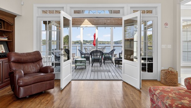 Une porte ouverte donne sur une terrasse aménagée avec du mobilier d’extérieur, et à l’intérieur, on aperçoit un fauteuil en cuir marron, le tout offrant une vue sur le paysage.