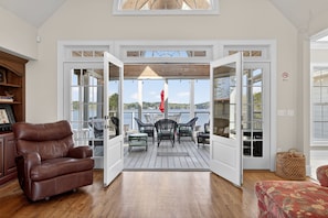 An open door leading to a deck with outdoor furniture, a brown leather chair inside, and a view of the landscape.