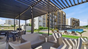 A covered outdoor seating area with wicker furniture, a pool, and modern buildings in the background. - Apartment in Bávaro Punta Cana | Swimming Pool | Near the Beach (Punta Cana)