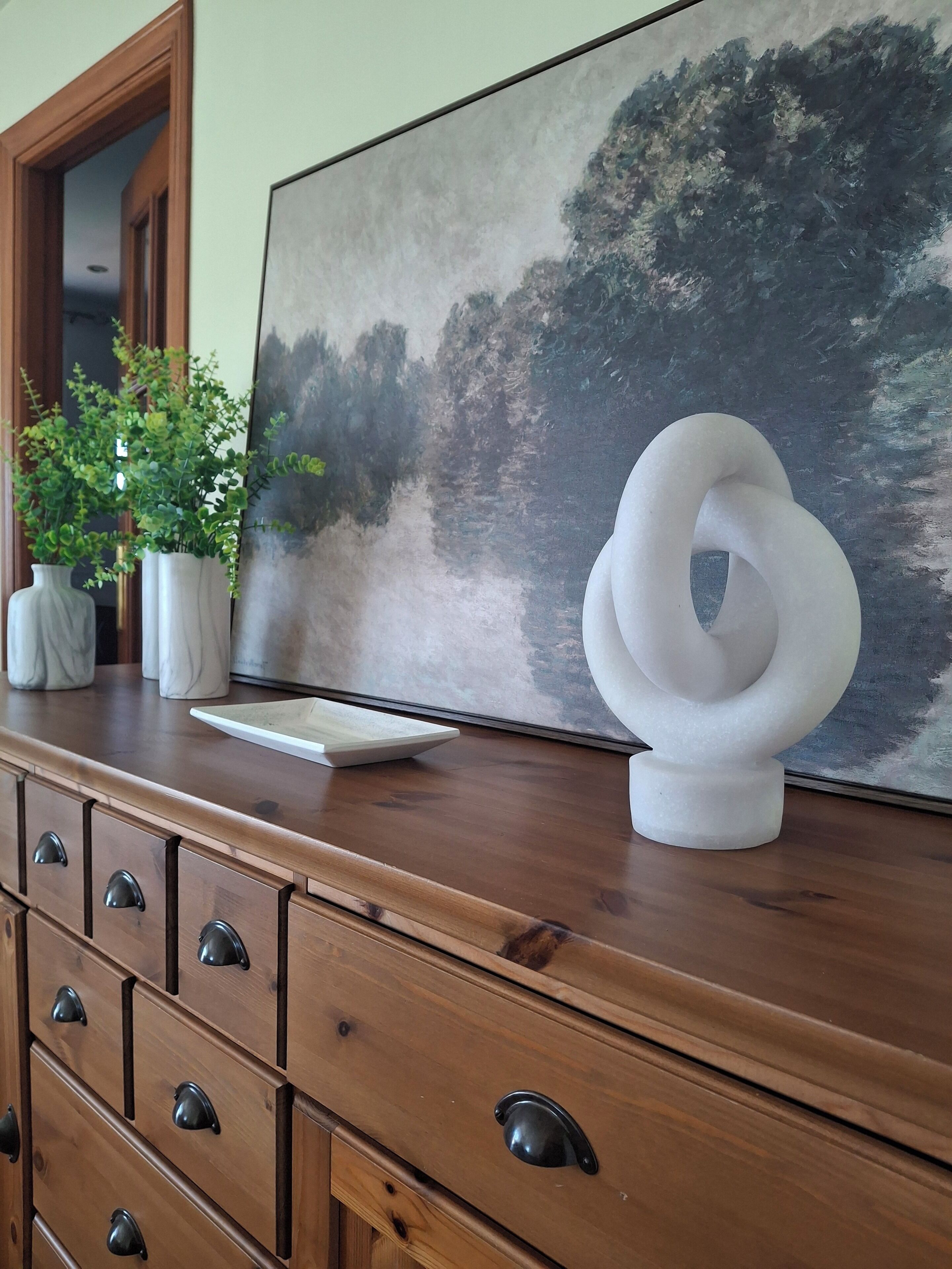Eine Holzkommode mit Schubladen, eine weiße abstrakte Skulptur und ein Baumgemälde.