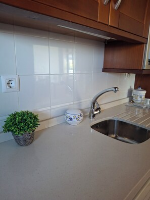 Uma cozinha com azulejos brancos, uma pia de aço inoxidável, uma planta em vaso e uma tigela decorativa.