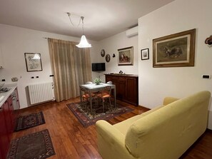 Uma sala de estar com um sofá amarelo, uma mesa de jantar com cadeiras, uma cozinha com radiador e uma televisão na parede.