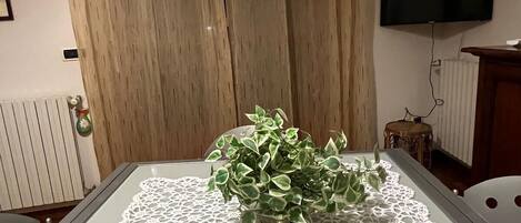 Una habitación con una mesa cubierta con un mantel de encaje, una planta en maceta, un televisor montado en la pared y un radiador.