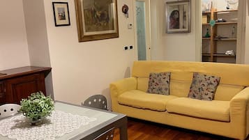 A living room with a yellow sofa, a dining table with chairs, a patterned rug, and a ceiling light fixture.