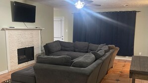 Ein Wohnzimmer mit Kamin, einem großen grauen Sofa, einem Deckenventilator und einem an der Wand montierten Fernseher.