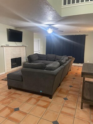 Una sala de estar con chimenea, un sofá grande de color gris, un ventilador de techo y un televisor montado en la pared.