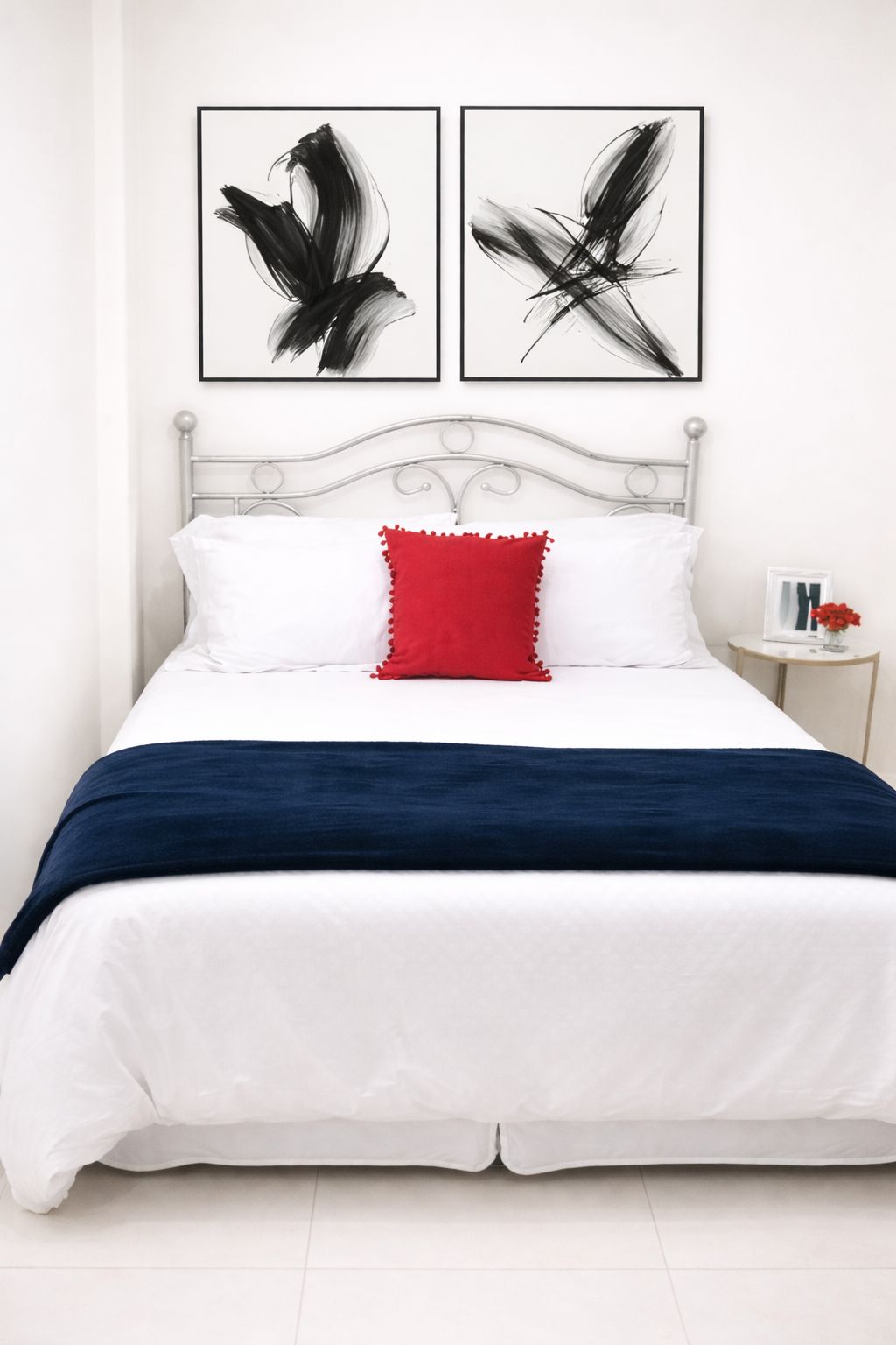 Una cama con ropa de cama blanca, un almohárraga rojo y dos obras de arte abstracto encima.