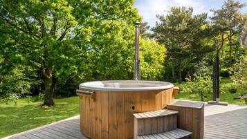 Un bain à remous en bois installé sur une terrasse avec des marches, entouré d’une végétation luxuriante.