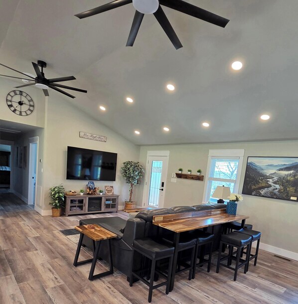 A spacious living area with a ceiling fan, recessed lighting, a flat-screen TV, a wooden dining table with chairs, a sofa, and a large window with a view of a river.