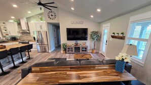 Uma cozinha moderna com eletrodomésticos de aço inoxidável, uma área de jantar com mesa de madeira e cadeiras pretas, e uma sala de estar com sofá e televisão.