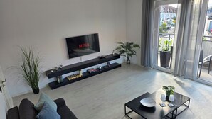 A modern living room with a flat-screen TV, a sofa, a coffee table, and a balcony with potted plants. - Modern Apartment with Balcony near Airport (Siegburg)