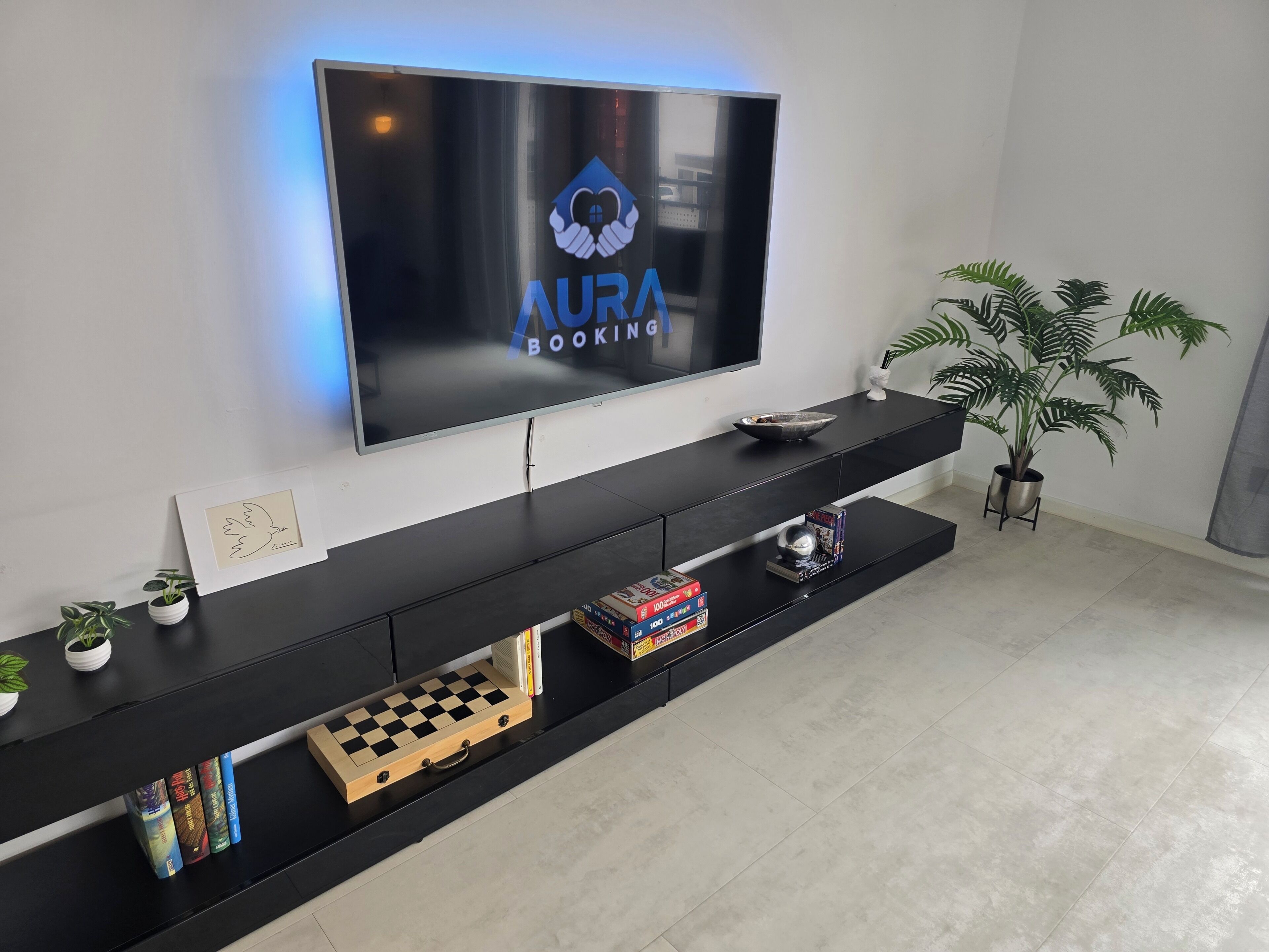 A modern living room with a flat-screen TV displaying 'AURA BOOKING' logo, a black entertainment unit with books and decorative items, and a potted plant.