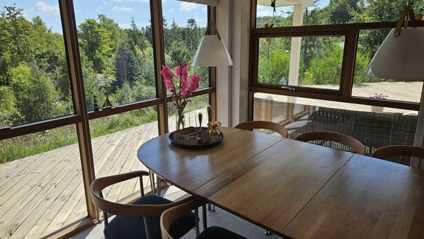 A wooden dining table with chairs, large windows, and a view of a forest. - 4 Star Holiday Home in Ebeltoft (Ebeltoft)