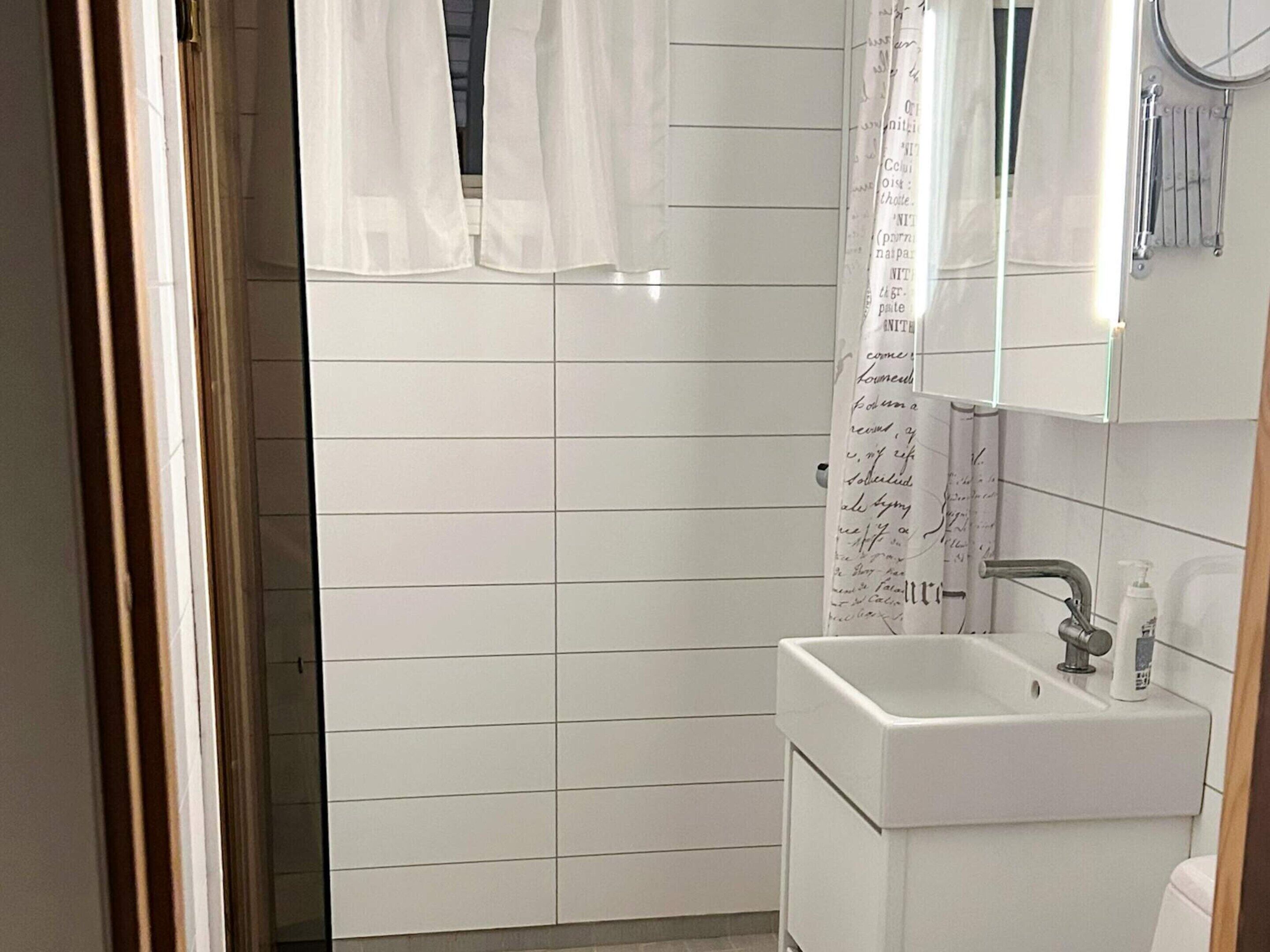 A bathroom with white tiles, a sink, a mirror, and a shower curtain with text.