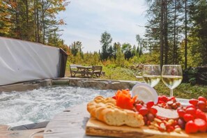 A hot tub with a wooden board of bread and grapes, two glasses of white wine, and a tent in a forest setting. - Chalet 3M & Spa - Lanaudiere (Chertsey)